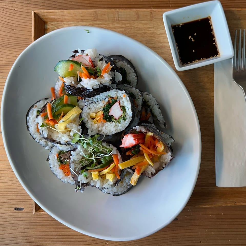 A white plate of Korean kimbap rolls with various fillings, next to a soy sauce dish.
