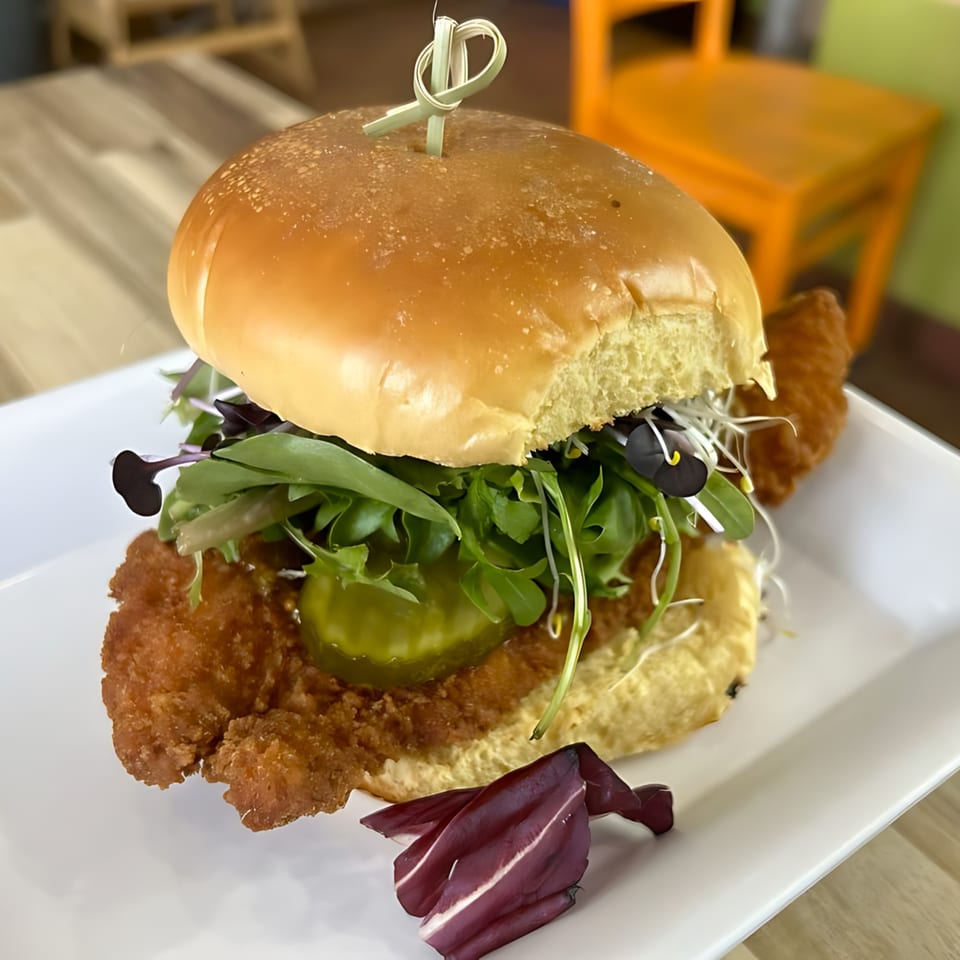 A Korean fried chicken sandwich with pickles, greens, and sprouts on a white plate.